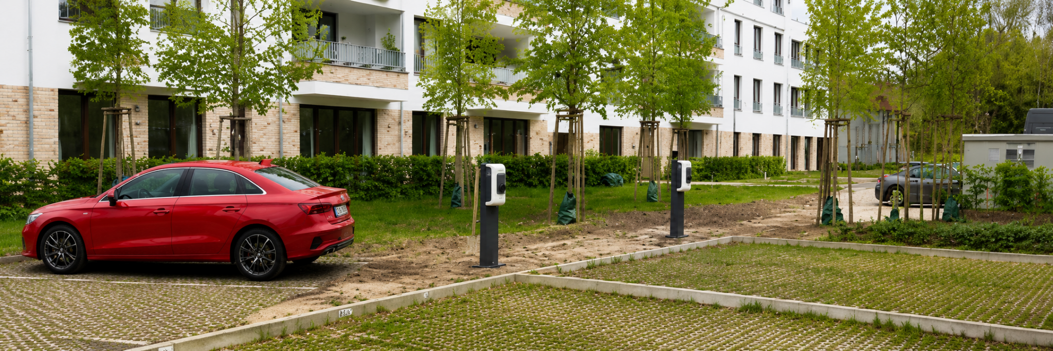 Vor einem Mehrfamilienhaus stehen auf einem Parkplatz mehrere Ladesäulen und ein rotes Auto